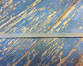 Può includere: Una barra metallica argentata, lunga e sottile. La barra è rettangolare con bordi arrotondati. Lo sfondo è una superficie di legno blu invecchiato con usura visibile, che aggiunge un'estetica rustica.