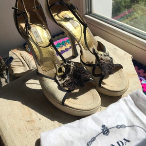 May include: A pair of beige platform sandals with black straps and a black and brown feather detail. The sandals have a high heel and are made by Prada.