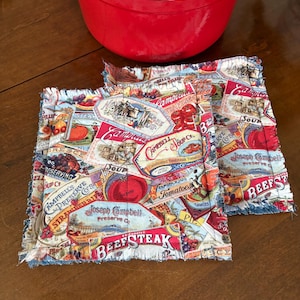 May include: Two square fabric pot holders with a frayed edge, featuring a vintage Campbell's Soup label design. The labels include images of tomatoes, strawberries, and beefsteak. A red pot is in the background.