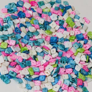 May include: A pile of small, square-shaped beads in various colors. The beads are primarily pink, white, blue, and green, with a slightly iridescent sheen. The beads appear to be made of a shell-like material, and are scattered across a white surface.