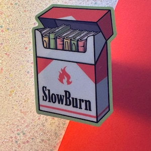 Puede incluir: Una pegatina con un dise&ntilde;o estilizado de un paquete de cigarrillos con la frase "SlowBurn" y un gr&aacute;fico de llama. El paquete es blanco con detalles rojos y contiene cigarrillos. La pegatina tiene un borde verde.