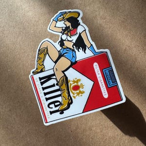 Op de afbeelding: Een sticker met een cowgirl in een witte top, blauwe shorts en cowboylaarzen, bovenop een sigarettenpakje. Het pakje is rood en wit met het woord "Killer" in vette zwarte letters. Retro design.