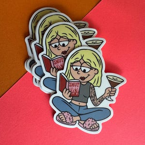May include: A stack of stickers featuring a cartoon woman with blonde hair, glasses, and a tattoo, reading a book titled "SMUT" with flames. She holds a cocktail and wears a gray top, blue jeans, and pink slippers. The stickers are on a pink and orange background.