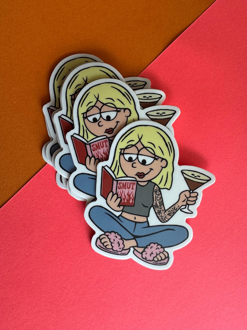 May include: A stack of stickers featuring a cartoon woman with blonde hair, glasses, and a tattoo, reading a book titled "SMUT" with flames. She holds a cocktail and wears a gray top, blue jeans, and pink slippers. The stickers are on a pink and orange background.