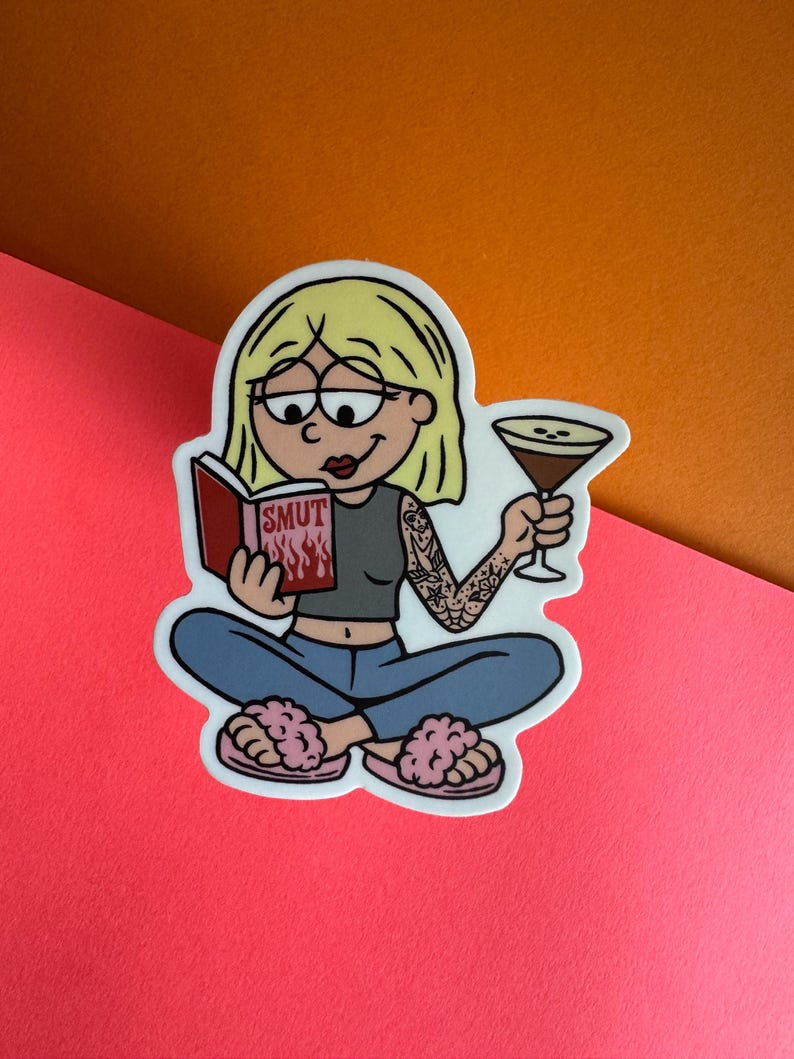 May include: A sticker featuring a cartoon illustration of a person with blonde hair, reading a book titled "SMUT" with flames, and holding a cocktail. The person is wearing a gray top, blue jeans, and pink fuzzy slippers. The person has a tattoo on their arm.
