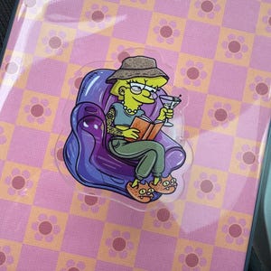 May include: A clear sticker featuring a cartoon character with a tattoo, reading a book, and holding a martini. The sticker is on a pink and orange floral patterned background. The character is wearing a hat, glasses, and a necklace.
