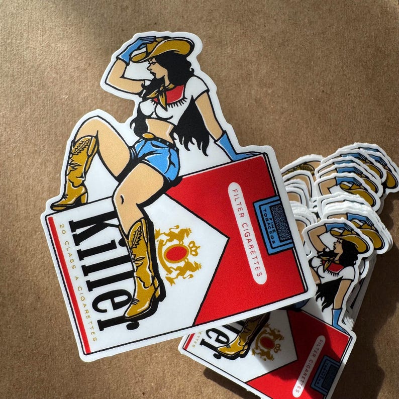 Op de afbeelding: Een sticker met een illustratie van een cowgirl bovenop een rood-witte sigarettenverpakking. De cowgirl draagt een gele hoed, een blauwe korte broek en cowboylaarzen. Het woord "Killer" staat op de verpakking.