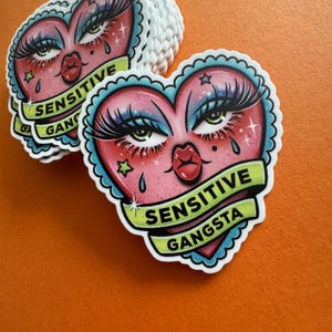 Op de afbeelding: Hartvormige stickers met een cartoongezicht. Het gezicht heeft lange wimpers, blauwe oogschaduw en een rode mond. De tekst "SENSITIVE GANGSTA" staat op een groene banner.