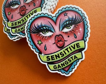 May include: A stack of heart-shaped stickers with a cartoon face design. The face has long eyelashes, blue eyeshadow, and a red pout. The text "SENSITIVE GANGSTA" is on a green banner across the heart.