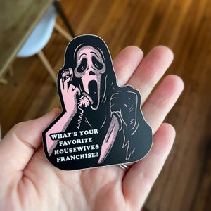 Op de afbeelding: Een zwart-witte sticker van Ghostface uit de Scream-franchise die een telefoon en een mes vasthoudt. De sticker heeft roze tekst die luidt "Wat is je favoriete housewives-franchise?"