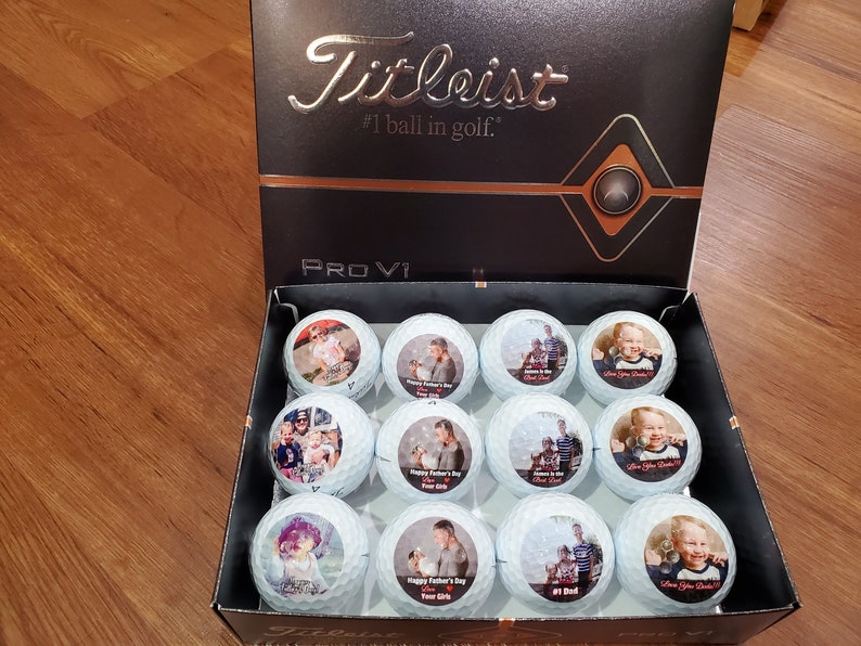 May include: A box of white golf balls with personalised photos printed on them. The box is black with the words "Titleist #1 ball in golf." and "PROV1" printed on it.