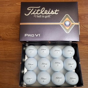 May include: A box containing twelve white Titleist Pro V1 golf balls. The box is open and the balls are arranged in three rows of four. Each ball has the Titleist logo and the number 1, 2, 3, or 4 printed on it.