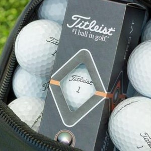 May include: A black golf bag with a zipper open, revealing a box of Titleist golf balls. The box is labelled "Titleist #1 ball in golf." The bag is filled with white golf balls.