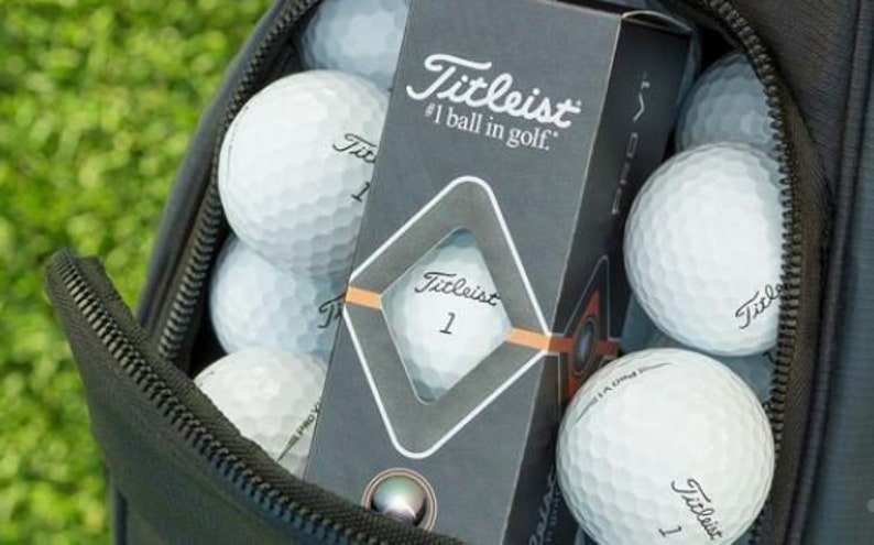 May include: A black golf bag with a zipper open, revealing a box of Titleist golf balls. The box is labelled "Titleist #1 ball in golf." The bag is filled with white golf balls.