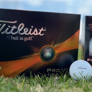 May include: A box of Titleist Pro V1 golf balls with a single golf ball in the foreground. The box is black with orange and silver accents and the text "Titleist #1 ball in golf." The golf ball is white with the Titleist logo and the number 2.