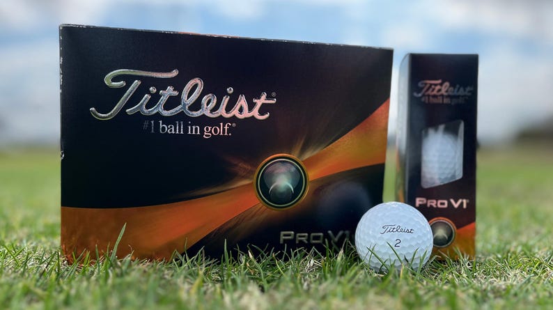 May include: A box of Titleist Pro V1 golf balls with a single golf ball in the foreground. The box is black with orange and silver accents and the text "Titleist #1 ball in golf." The golf ball is white with the Titleist logo and the number 2.