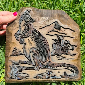 May include: A rectangular wooden printing block featuring a deeply carved relief of a kangaroo standing in a desert landscape with mountains and small plants. The wood has a dark brown finish with visible grain and carved lines throughout.