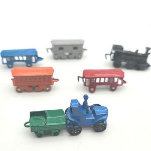 May include: A collection of miniature toy trains and train cars in various colors, including blue, red, orange, green, gray, and black. The toys are made of metal and are arranged on a white surface.
