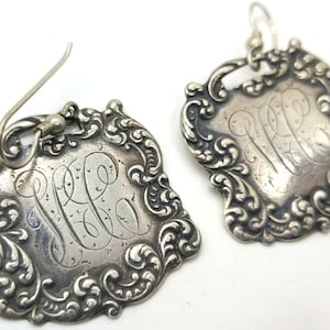 May include: A pair of silver earrings with a monogrammed design. The earrings are shaped like a square with ornate details around the edges. The monogram is in the center of the square and is surrounded by a decorative border.