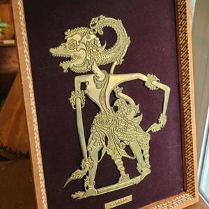 May include: A gold-colored metal shadow puppet of a mythical figure, framed in a brown wood frame with a burgundy velvet backing. The puppet is in a dancing pose with arms outstretched and the text "Wayang" is printed below the figure.
