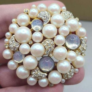 May include: A round brooch featuring a cluster of faux pearls in varying sizes, interspersed with iridescent, light blue cabochons and small clusters of clear rhinestones. The brooch has a gold-tone metal setting.