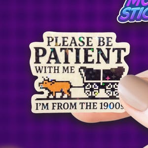 May include: A sticker with the text "PLEASE BE PATIENT WITH ME I'M FROM THE 1900s" and a pixelated image of a cow pulling a cart. The sticker is held against a purple background with smiley faces and stars.