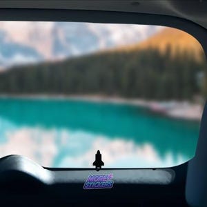 May include: A black sticker in the shape of a spaceship is affixed to a car window. The sticker is in front of a blurred scenic view of a lake and mountains. The text "MORE STICKERS" is visible on the bottom right.