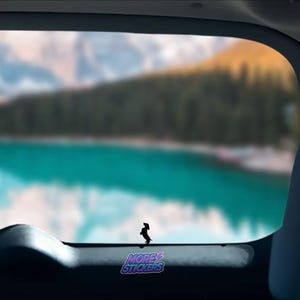 May include: A black silhouette of a dachshund dog sticker on a car window with a blurred background of a lake and mountains. The sticker says "More Stickers" in a purple and blue gradient.