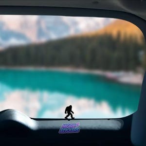 May include: A black silhouette of Bigfoot walking across a car window with a blue lake and mountains in the background. The sticker says "More Stickers" in purple and white text.