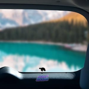 May include: A black silhouette of a turtle sticker on a car window with a blurred background of a lake and mountains. The sticker also includes the text "MORE STICKERS" in a purple and blue gradient.