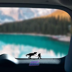 May include: A black silhouette of a Bigfoot walking a Tyrannosaurus Rex on a leash. The image is a sticker placed on the back window of a car. The background is a blurry image of a lake and mountains.