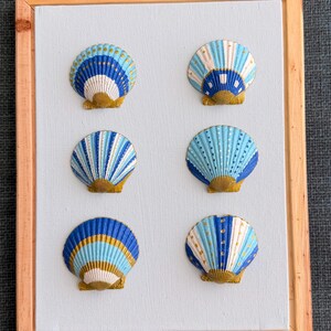 May include: A framed artwork featuring six painted seashells. The shells are in shades of blue, white, and gold, with various patterns. The frame is made of light brown wood, and the background is a light blue canvas.
