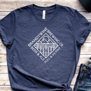 May include: A navy blue t-shirt with a white graphic of a logo for Brandywyne Brewing Co. The logo features a stylized image of a landscape with a sun, hills, and a fence. The text around the image reads "Brandywine Brewing Co. Beer & Ale Full & 1/2 Pint."