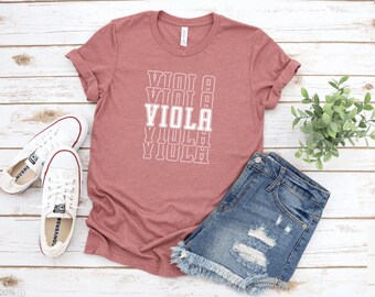 May include: A mauve t-shirt with the word "VIOLA" repeated in white text in a stacked, retro design. The shirt is laid flat on a white wooden surface with a pair of white sneakers and denim shorts.