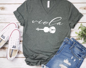 May include: A dark green V-neck t-shirt with the word "viola" written in white cursive script and a white outline of a viola underneath. The shirt is laying on a white wooden surface with a pair of white sneakers and a pair of blue denim shorts.