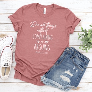 May include: A maroon t-shirt with the text "Do all things without complaining or arguing Philippians 2:14" in white. The shirt is laying on a wooden surface with a pair of white sneakers and a pair of blue denim shorts.