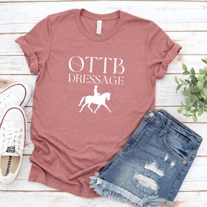 May include: A maroon t-shirt with white text that reads "OTTB Dressage" and a white silhouette of a rider on a horse.