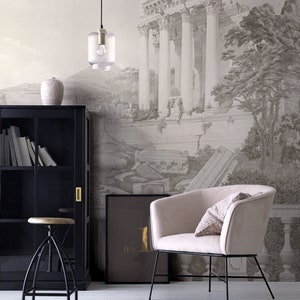 Op de afbeelding: Een woonkamerinterieur met een zwarte boekenkast, een houten kruk, een lamp, een beige fauteuil met een gepatroneerd kussen en een ingelijst drukwerk met de tekst "no. 1" op een muur met een behang dat een zwart-witte tekening van een oude Romeinse ru&iuml;ne voorstelt.