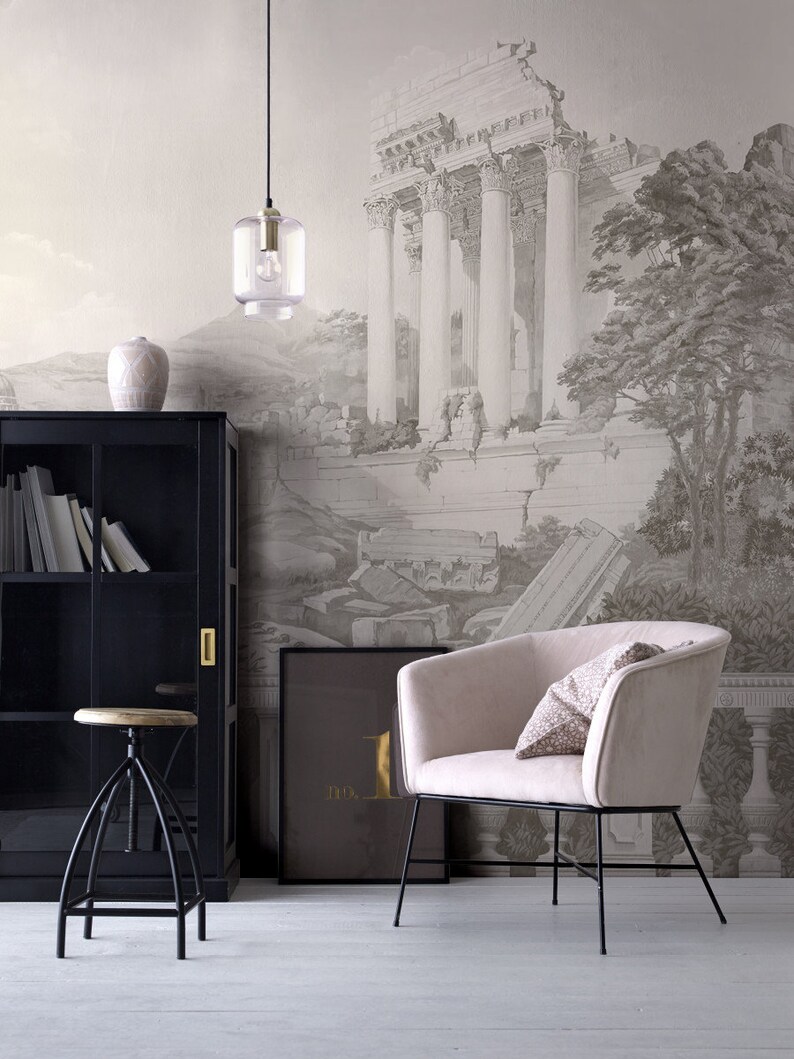 Op de afbeelding: Een woonkamerinterieur met een zwarte boekenkast, een houten kruk, een lamp, een beige fauteuil met een gepatroneerd kussen en een ingelijst drukwerk met de tekst "no. 1" op een muur met een behang dat een zwart-witte tekening van een oude Romeinse ru&iuml;ne voorstelt.