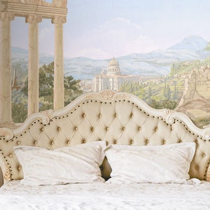 Op de afbeelding: Een wit gestoffeerd bed met een knoopsteun, twee witte kussens en een wit dekbed. Het bed staat voor een muur met een muurschildering die een landschap met ru&iuml;nes en kolommen voorstelt.