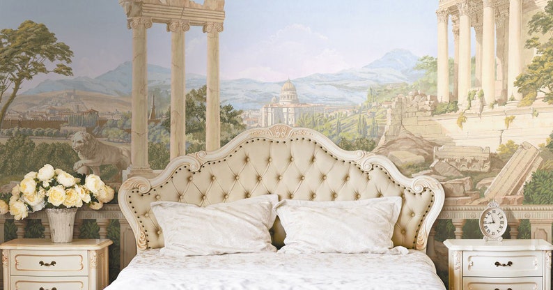 Op de afbeelding: Een wit gestoffeerd bed met een knoopsteun, twee witte kussens en een wit dekbed. Het bed staat voor een muur met een muurschildering die een landschap met ru&iuml;nes en kolommen voorstelt.