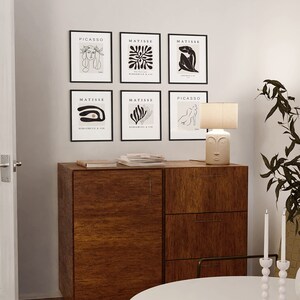 May include: Six black framed art prints with white line drawings of people and abstract shapes. The prints are arranged in two rows of three. The top row reads "PICASSO", "MATISSE", "MATISSE". The bottom row reads "MATISSE", "MATISSE", "PICASSO". A wooden cabinet with three drawers is below the prints. A white round table with a white ceramic candle holder with three white candles is in the foreground.