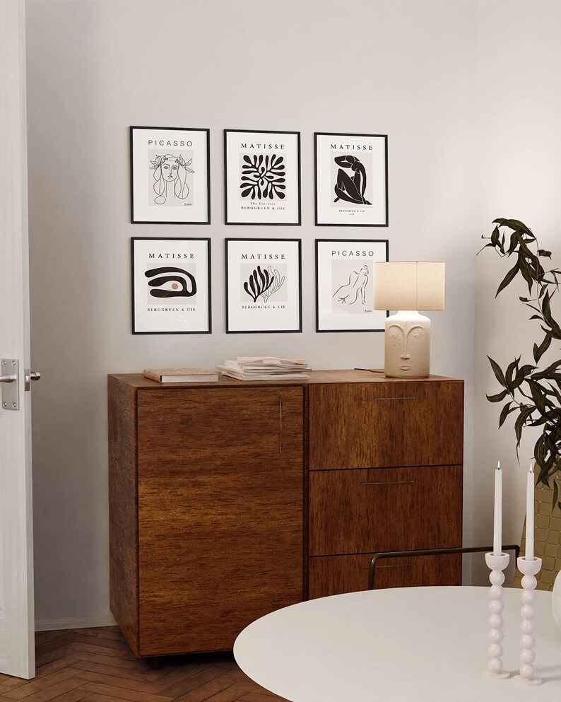 May include: Six black framed art prints with white line drawings of people and abstract shapes. The prints are arranged in two rows of three. The top row reads "PICASSO", "MATISSE", "MATISSE". The bottom row reads "MATISSE", "MATISSE", "PICASSO". A wooden cabinet with three drawers is below the prints. A white round table with a white ceramic candle holder with three white candles is in the foreground.