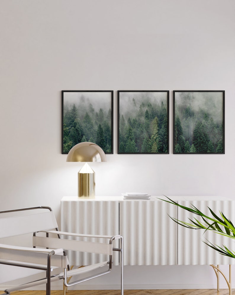 May include: Three black framed prints of a foggy forest scene. The prints are hung on a white wall above a white cabinet with a gold lamp and a white chair.