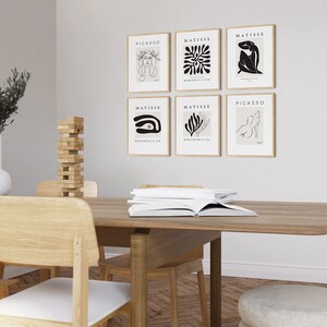 May include: Six framed art prints featuring abstract line drawings and the names of artists Picasso and Matisse. The prints are hung on a white wall above a wooden table with chairs.