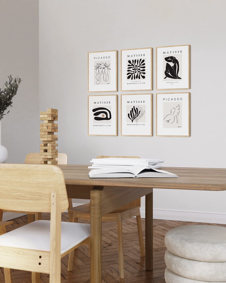 May include: Six framed art prints featuring abstract line drawings and the names of artists Picasso and Matisse. The prints are hung on a white wall above a wooden table with chairs.