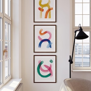 May include: Three framed abstract art prints with colorful brushstrokes in yellow, orange, red, pink, blue, and green. The prints are arranged vertically on a white wall.