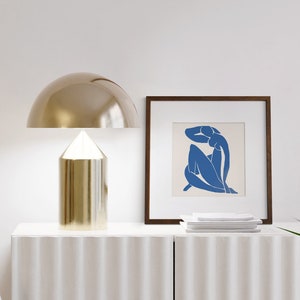 May include: A gold table lamp with a dome-shaped shade sits on a white dresser with a white framed print of a blue silhouette of a woman. The print is on a white background.
