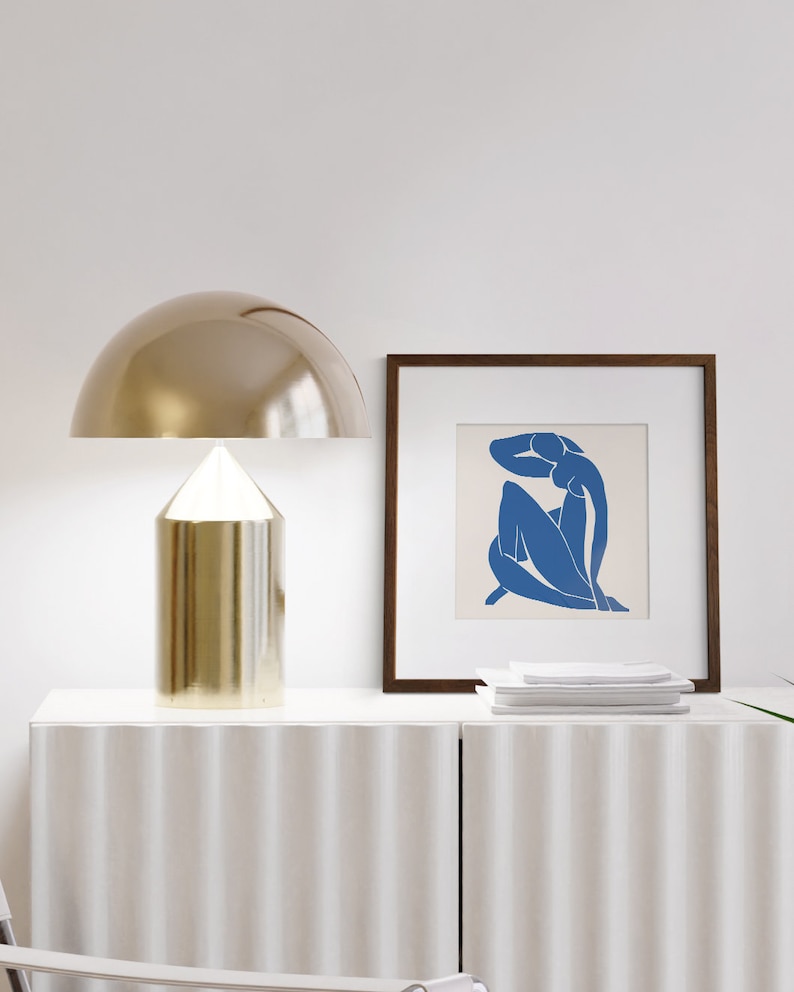 May include: A gold table lamp with a dome-shaped shade sits on a white dresser with a white framed print of a blue silhouette of a woman. The print is on a white background.
