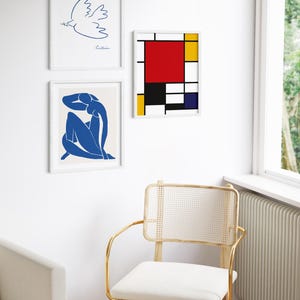 May include: Three framed art prints on a white wall. The top print is a blue line drawing of a dove with an olive branch in its beak. The middle print is a blue silhouette of a seated woman. The bottom print is a geometric abstract painting with red, yellow, blue, and black squares.